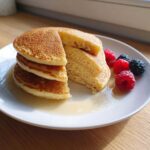 A stack of fluffy almond flour pancakes, cut to show the texture, served with maple syrup and fresh raspberries and blackberries.