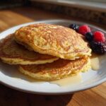 A stack of three golden brown almond flour pancakes drizzled with syrup and topped with fresh raspberries, blueberries, and blackberries.