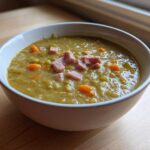 A close-up of a white bowl filled with thick, green pea soup, topped with diced ham and carrots.