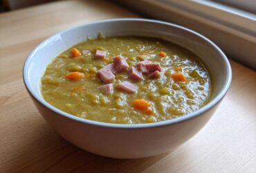 A close-up of a white bowl filled with thick, green pea soup, topped with diced ham and carrots.