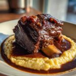 A single, dark, glossy braised short ribs bone-in portion served atop creamy polenta, drenched in rich brown sauce.