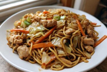 A close-up of a plate piled high with homemade chicken chow mein recipe, featuring noodles, chicken pieces, carrots, and cabbage.