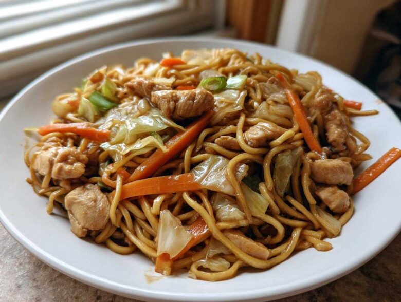 A close-up of a plate piled high with homemade chicken chow mein recipe, featuring noodles, chicken pieces, carrots, and cabbage.