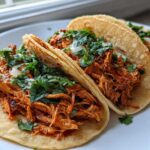 Close-up of three soft corn tortilla chicken tacos filled with shredded, saucy chicken, topped with fresh cilantro and onion.