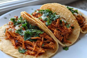 Close-up of three soft corn tortilla chicken tacos filled with shredded, saucy chicken, topped with fresh cilantro and onion.