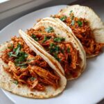Close-up of three soft tortilla chicken tacos filled with shredded, saucy chicken and topped with fresh cilantro.