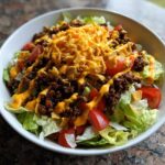 A close-up view of an amazing taco salad featuring lettuce, ground beef, tomatoes, cheese, and a drizzle of cheese sauce.