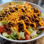 A close-up view of an amazing taco salad featuring seasoned ground beef, shredded cheese, tomatoes, and dressing over lettuce in a white bowl.