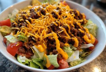 A close-up view of an amazing taco salad featuring seasoned ground beef, shredded cheese, tomatoes, and dressing over lettuce in a white bowl.
