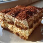 A close-up of a perfect slice of tiramisu recipe, showing layers of cream and coffee-soaked ladyfingers, dusted with cocoa.