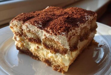 A close-up of a perfect slice of tiramisu recipe, showing layers of cream and coffee-soaked ladyfingers, dusted with cocoa.