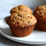 A close-up of a perfectly baked banana nut muffin topped with chopped walnuts on a white plate.