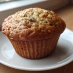 A close-up of a freshly baked, golden-brown banana protein muffin sitting on a small white saucer.