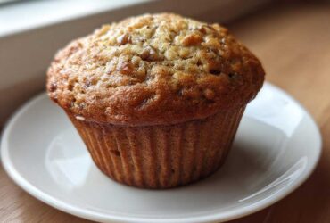 A close-up of a freshly baked, golden-brown banana protein muffin sitting on a small white saucer.