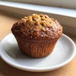 A close-up of a single, golden brown banana protein muffin topped with nuts and coarse sugar.