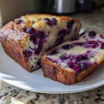 A freshly baked loaf of blueberry banana bread, sliced to show the moist interior packed with purple blueberries.