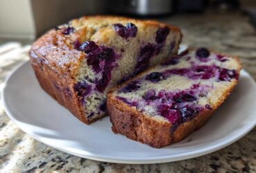 A freshly baked loaf of blueberry banana bread, sliced to show the moist interior packed with purple blueberries.