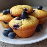 A stack of golden, freshly baked blueberry protein muffins garnished with fresh blueberries on a white plate.