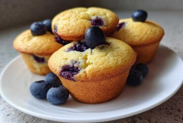 A stack of golden, freshly baked blueberry protein muffins garnished with fresh blueberries on a white plate.