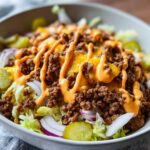 Close-up of a cheeseburger bowl featuring seasoned ground beef, melted cheese, special sauce drizzle, pickles, and red onion over lettuce.
