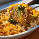 A spoonful of cheesy chicken and rice with melted cheddar cheese and broccoli florets being lifted from a white baking dish.