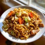 A close-up of a white plate filled with savory chicken chow mein recipe, featuring noodles, chicken pieces, carrots, and cabbage.