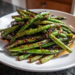 A plate piled high with blistered, glossy chinese green beans with garlic sauce, showing caramelized bits.