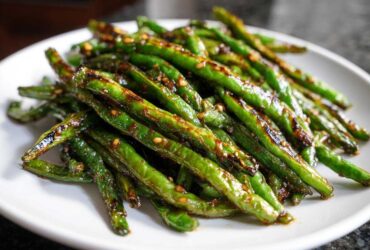 A pile of blistered, glossy chinese green beans with garlic sauce and sesame seeds on a white plate.
