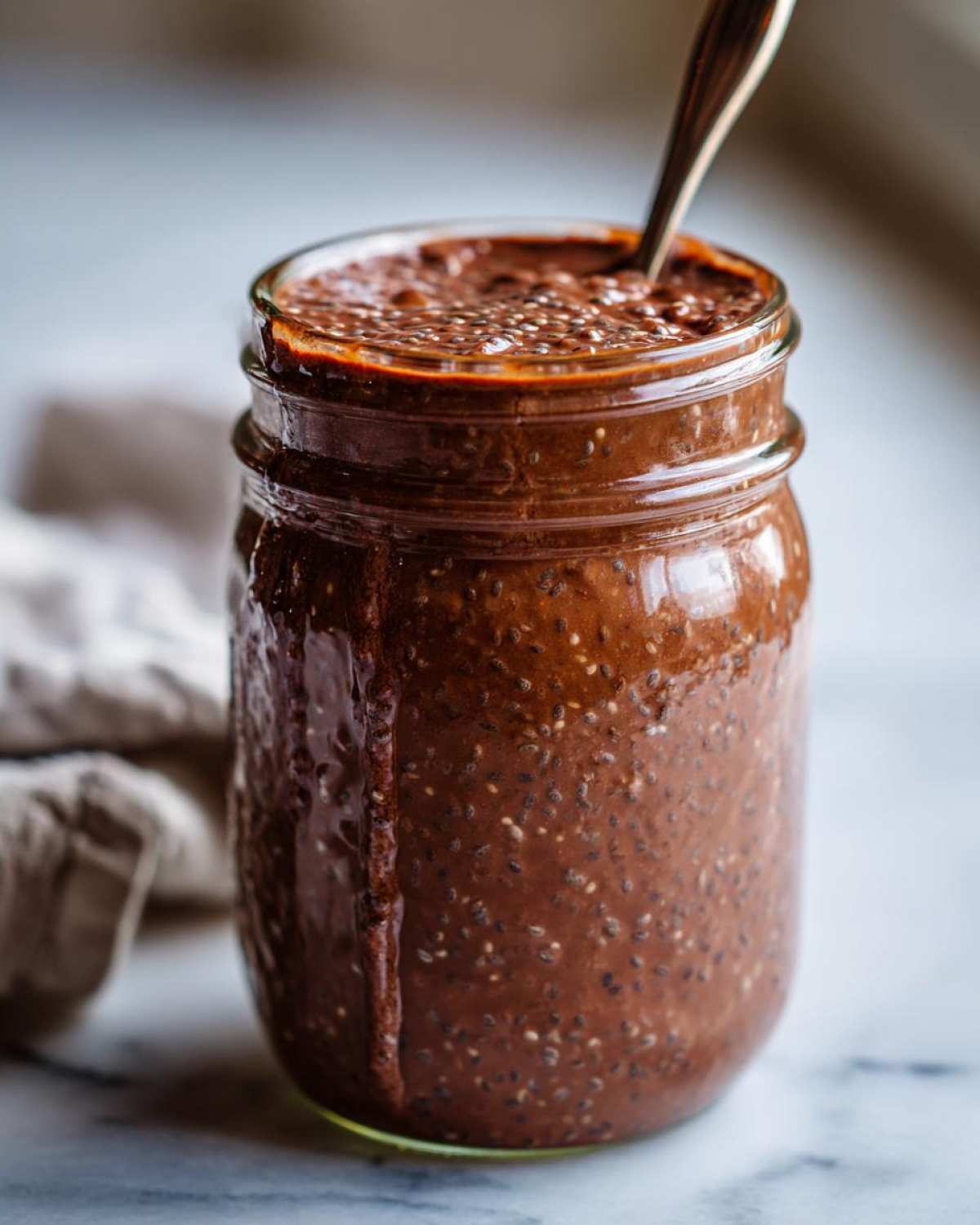 A close-up of rich, thick chocolate overnight oats with visible chia seeds in a glass jar, ready to eat.