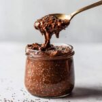 A spoonful of thick, rich chocolate overnight oats being lifted from a glass jar, showing chia seeds.
