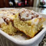 Close-up of a fluffy slice being lifted from a white baking dish of sweet Cinnamon roll casserole topped with icing.