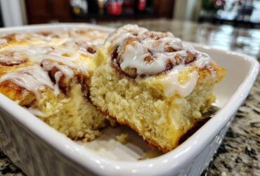 Close-up of a fluffy slice being lifted from a white baking dish of sweet Cinnamon roll casserole topped with icing.