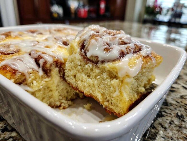 Close-up of a fluffy slice being lifted from a white baking dish of sweet Cinnamon roll casserole topped with icing.