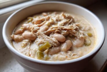 A close-up view of a white bowl filled with creamy crockpot white chicken chili, featuring shredded chicken, white beans, and green chiles.