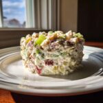 A perfectly molded serving of creamy ham salad with visible chunks of ham and celery, seasoned and placed on a white plate.