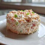 A perfectly molded serving of creamy ham salad with visible celery and red pepper pieces, served on a slice of bread.