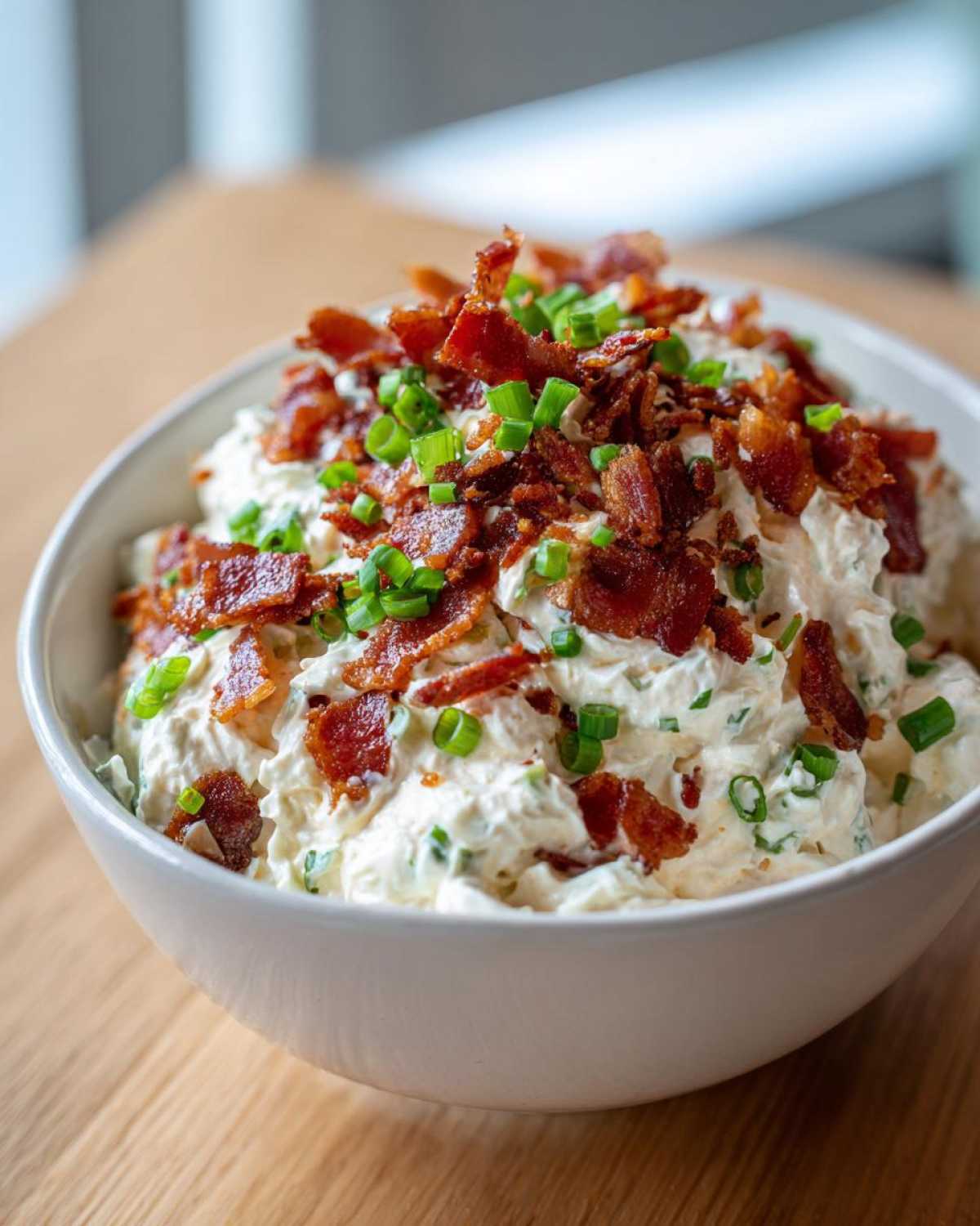 A white bowl filled with creamy Million Dollar Dip, generously topped with crispy bacon pieces and fresh green onions.