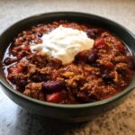 Close-up of a deep green bowl filled with rich, meaty crockpot chili topped with a dollop of sour cream.
