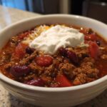 A close-up of a white bowl filled with rich crockpot chili, topped with sour cream and shredded cheese.