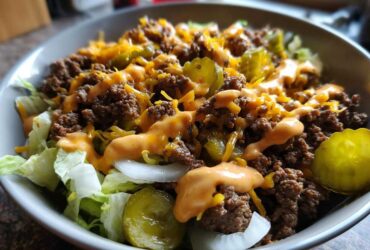 A close-up view of a hearty cheeseburger bowl featuring seasoned ground beef, shredded cheese, pickles, and special sauce over lettuce.