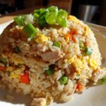 A mound of homemade chicken fried rice, featuring visible pieces of chicken, egg, peas, and carrots, topped with fresh green onions.