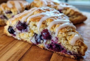 A close-up view of a moist, flaky blueberry scone drizzled with white icing, sitting on a wooden board.