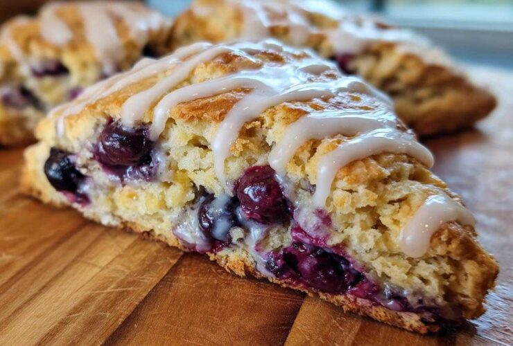 A close-up view of a moist, flaky blueberry scone drizzled with white icing, sitting on a wooden board.