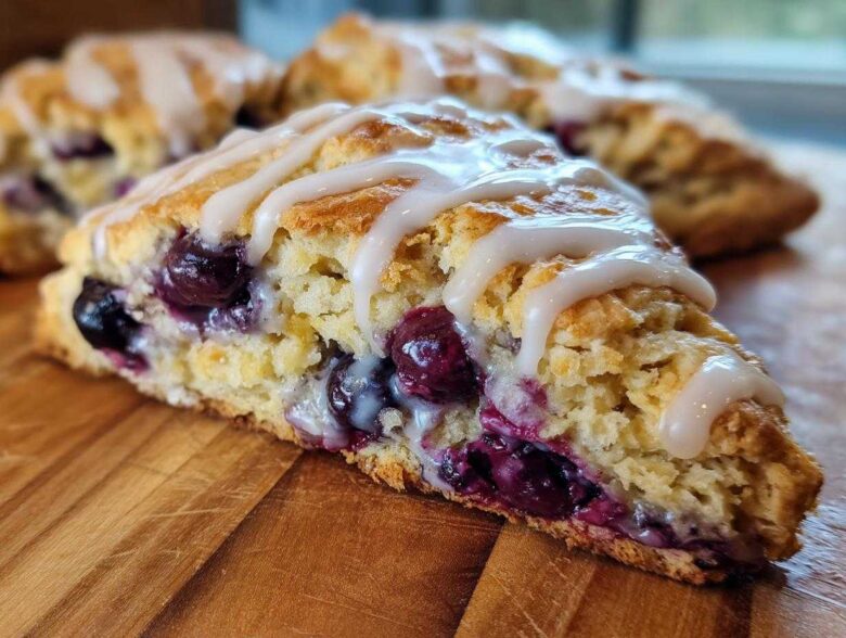 A close-up view of a moist, flaky blueberry scone drizzled with white icing, sitting on a wooden board.