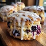 Close-up of a flaky blueberry scone topped with thick white glaze dripping down the sides, loaded with fresh blueberries.
