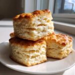 Three tall, flaky buttermilk biscuits with golden brown tops stacked on a white plate in bright sunlight.