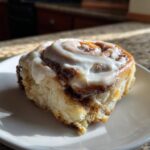 A single, freshly baked cinnamon roll topped generously with white icing, sitting on a white plate.