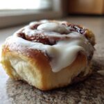 A close-up, low-angle shot of a single, freshly baked cinnamon roll topped with thick white icing.