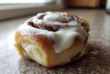 A close-up, low-angle shot of a single, freshly baked cinnamon roll topped with thick white icing.