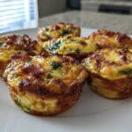 A close-up of six golden-brown Egg Muffin Cups topped with crispy bacon and visible spinach on a white plate.